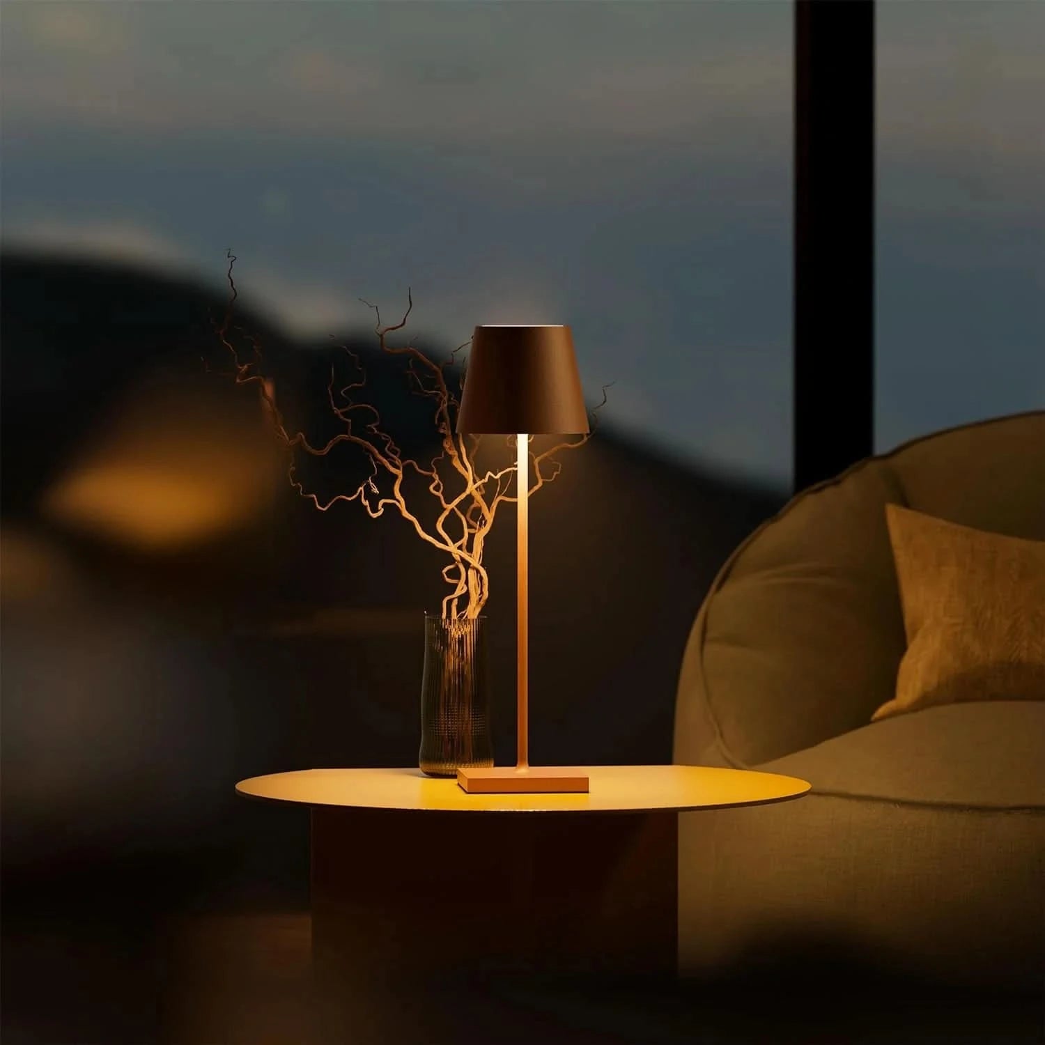 En stilren lampa med en brun lampskärm på ett runt bord bredvid en mysig stol, med kvistar i en vas.
