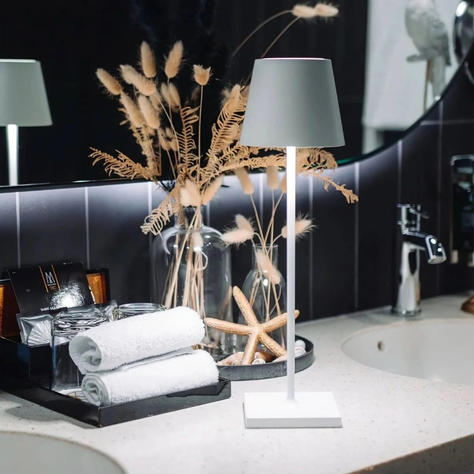 En modern vit lampa på ett badrumsbänk med dekorativa växter, handdukar och sjöstjärnor.